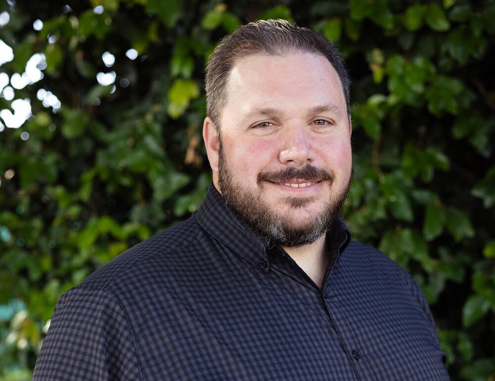 A man with a beard wearing a black checkered shirt, standing against a backdrop of green foliage.
