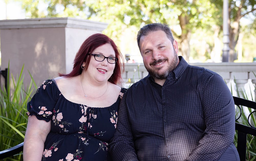 A smiling couple sits on a bench in a sunny park. The woman has red hair and glasses, wearing a floral dress; the man has short hair and a beard, wearing a dark checked shirt.