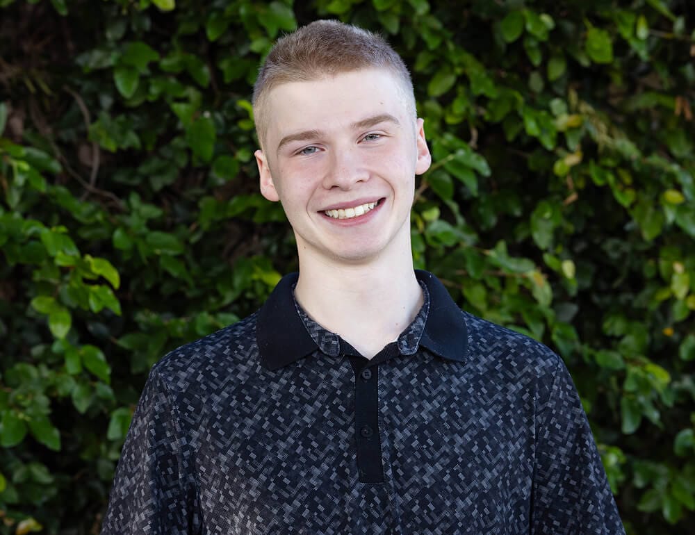Smiling young man in a patterned dark shirt stands in front of lush green foliage. The mood is cheerful and relaxed.