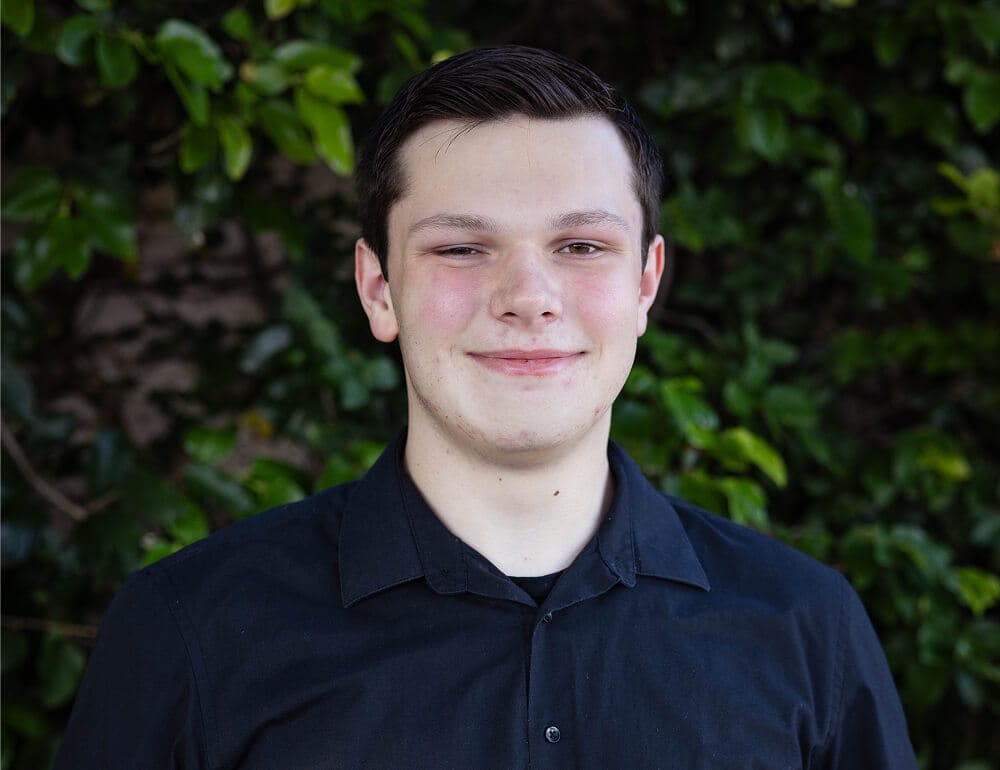 Young man smiling softly, wearing a black shirt, stands against a leafy green background, conveying a sense of calm and friendliness.