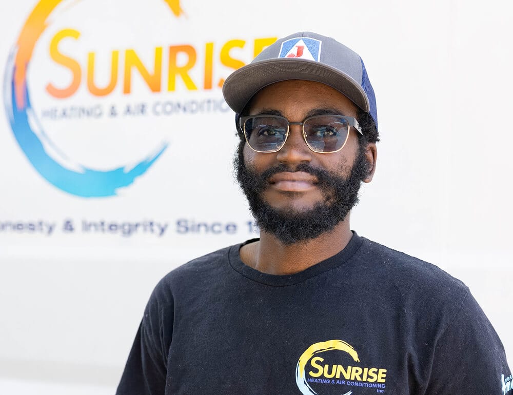 A man wearing glasses and a cap stands in front of a sign for Sunrise Heating & Air Conditioning. He has a beard and is wearing a black t-shirt with the company's logo.