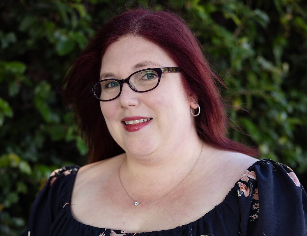A woman with red hair and glasses, wearing a floral dress.