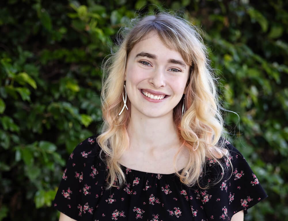 A young woman with wavy blonde hair and wearing a black floral dress, smiling in front of green foliage.